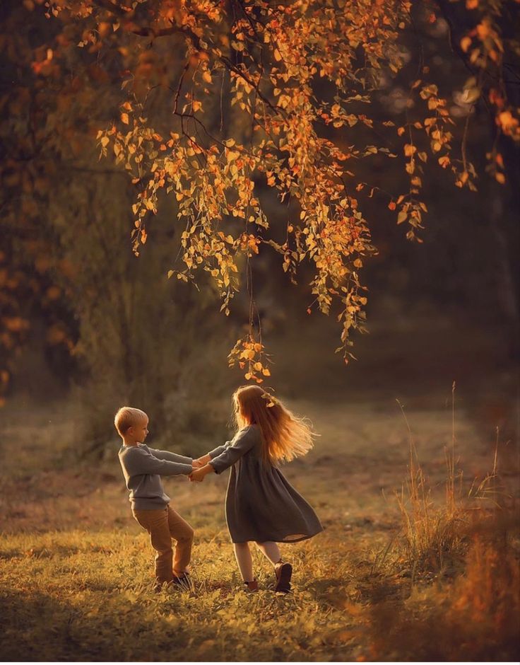 two young children holding hands under a tree in the fall time with leaves on it