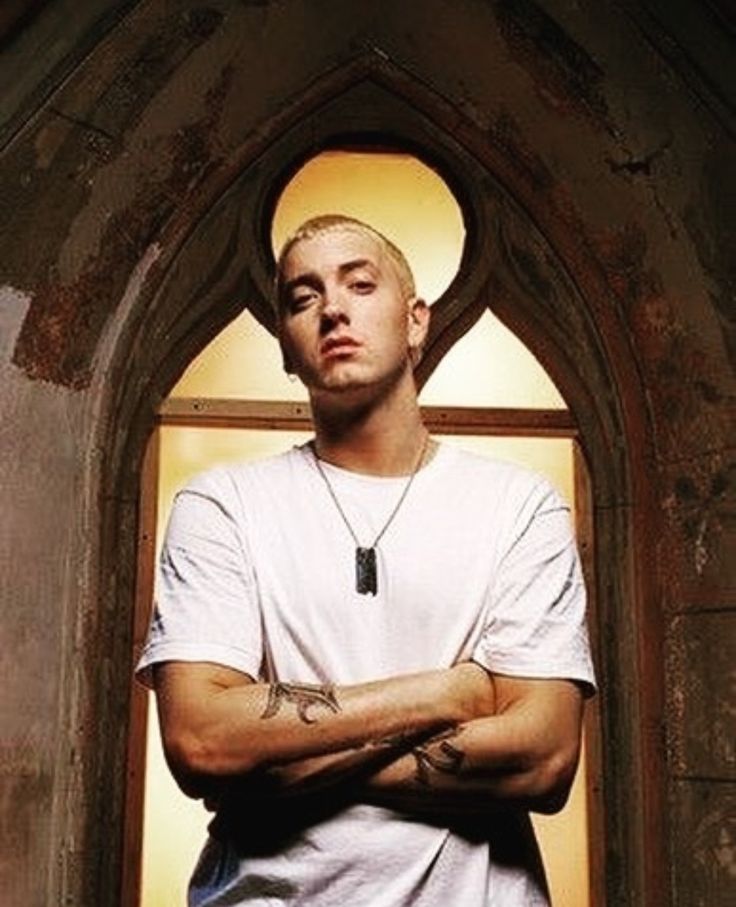 a man with his arms crossed standing in front of an arch doorway, wearing a white t - shirt and jeans