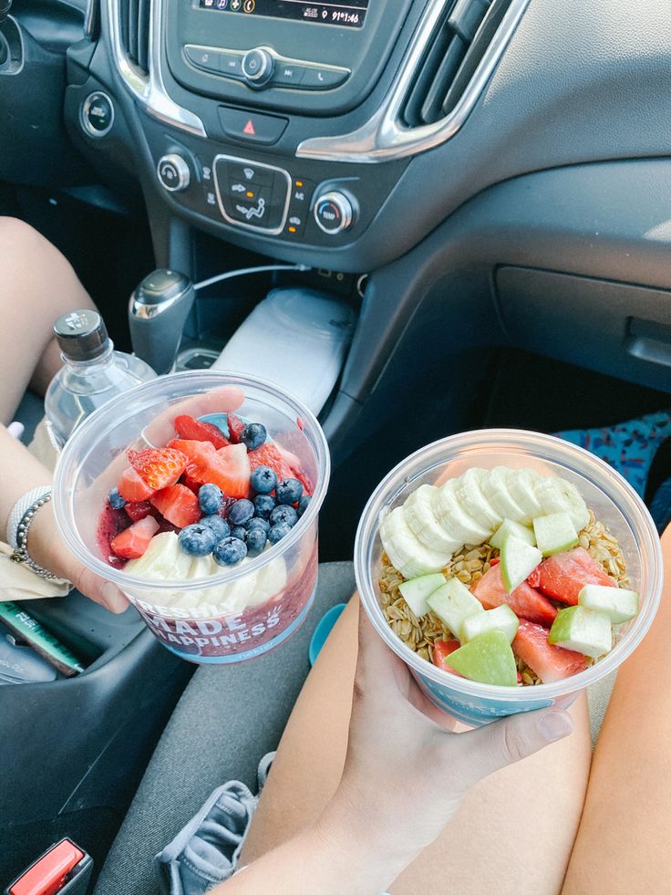 two people sitting in the drivers seat holding up bowls of fruit and yogurt