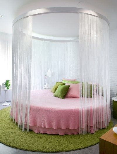 a round bed with sheer curtains on the top and green pillows on the bottom, in a white bedroom