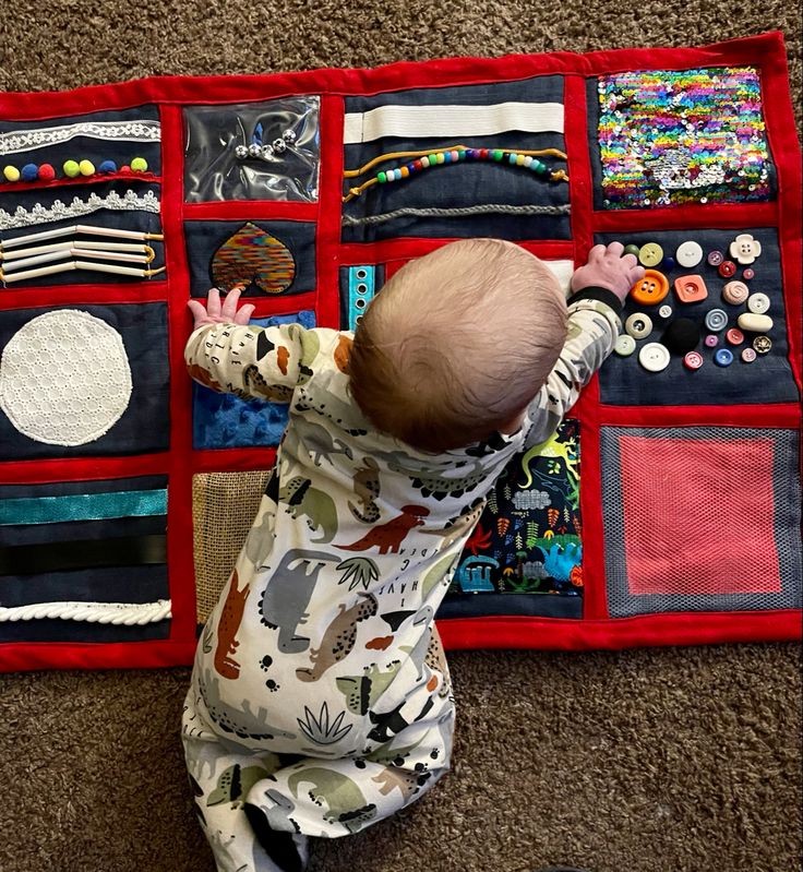 a baby standing on the floor in front of a quilted wall hanging with buttons