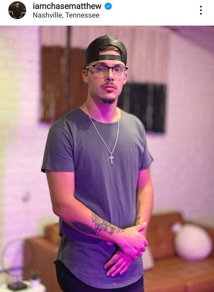 a man wearing glasses and a t - shirt standing in front of a couch with his arms crossed