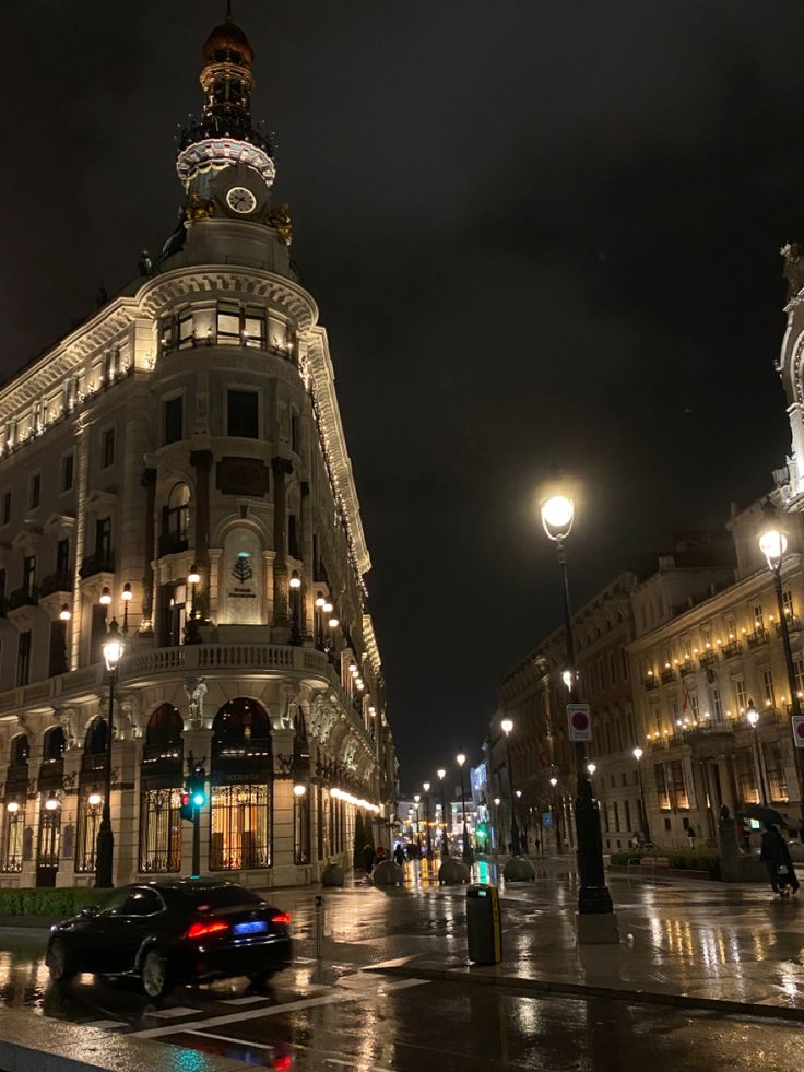 a very tall building sitting on the side of a wet street at night with traffic passing by