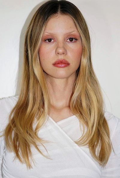 a woman with long blonde hair wearing a white shirt and looking at the camera while standing in front of a white wall