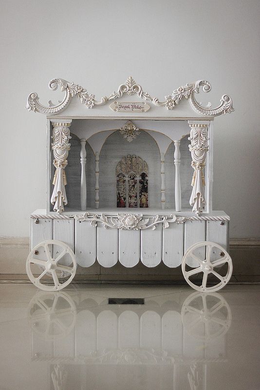 a white cart with wheels on the floor in front of a wall and mirror behind it