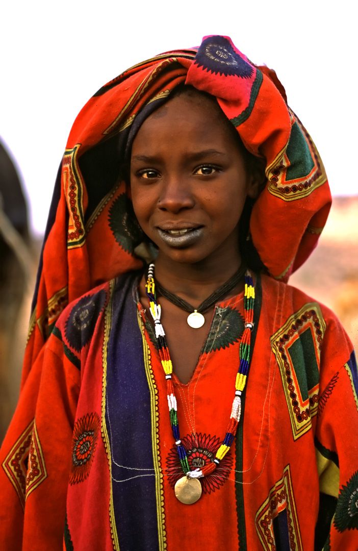 a young woman wearing an orange and black outfit with beads on her head, standing in front of a horse