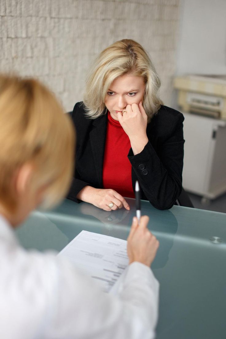 a woman sitting at a desk talking to another woman who is holding a pen in her hand