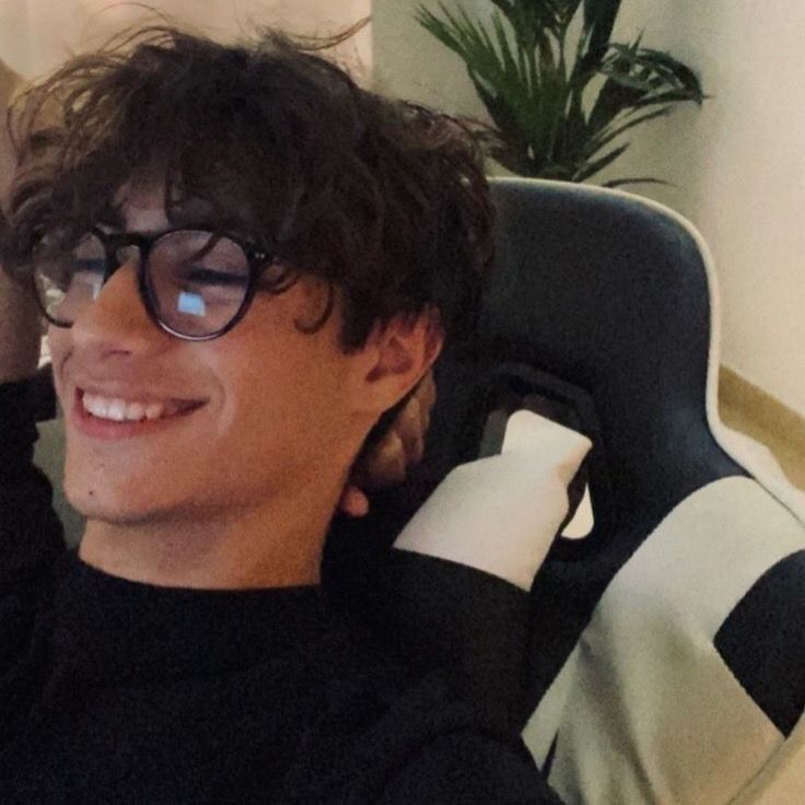 a young man wearing glasses sitting in a chair with his hand on his head and looking at the camera