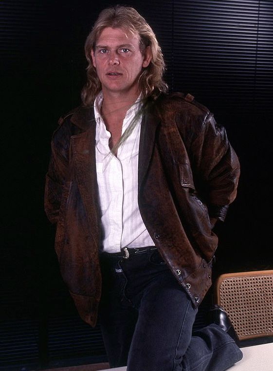 a man sitting on top of a white table next to a wooden chair and wearing a brown jacket