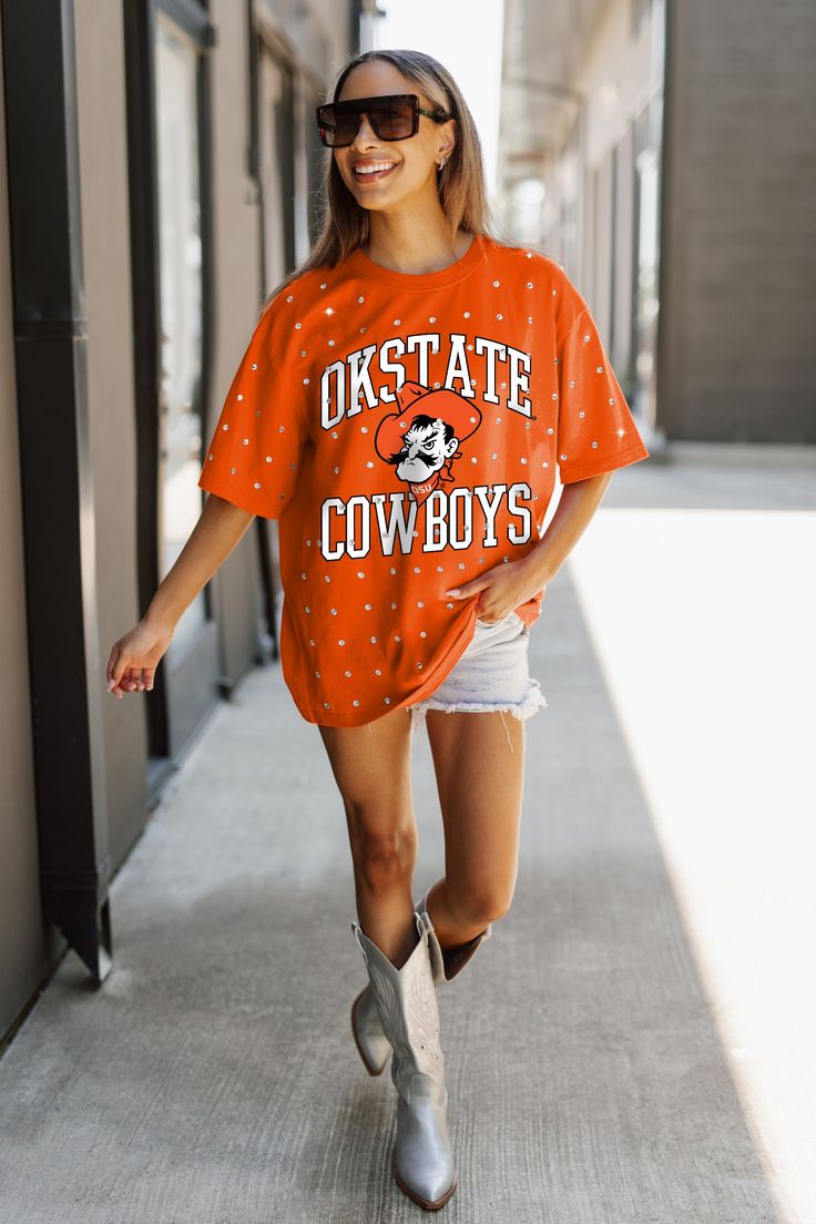 a woman in an orange shirt and cowboy boots is walking down the street with her hand on her hip