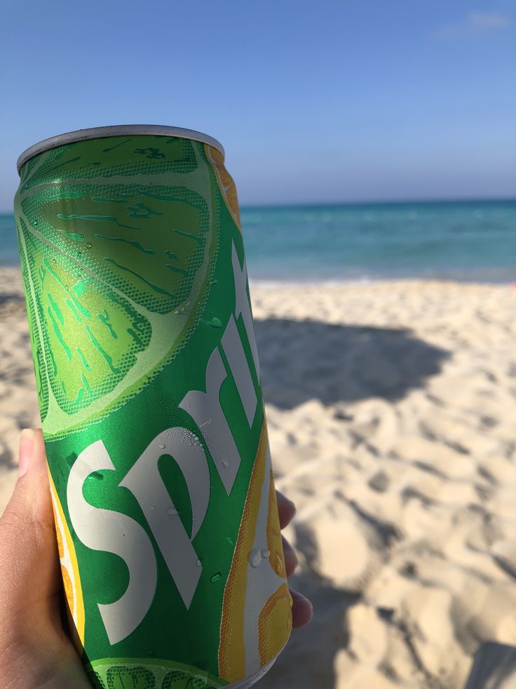 a hand holding up a can of spriter on the beach with water in the background