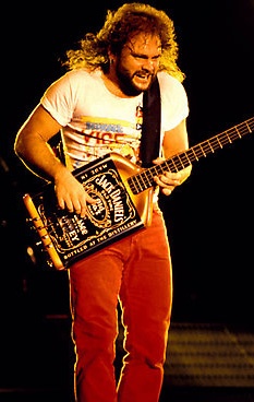 a man standing on stage with a guitar in his hand and wearing red pants, white t - shirt and black tie