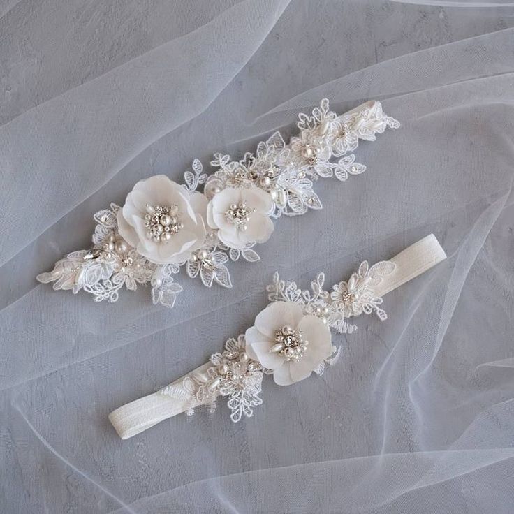 two bridal garters with white flowers and pearls on top of sheer fabric