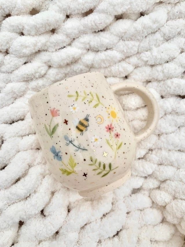 a white mug with flowers and bees on it sitting on top of a pile of blankets