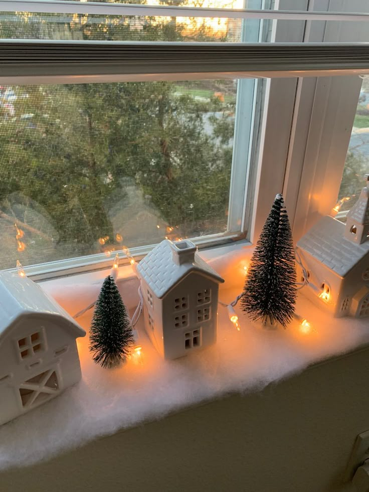 small christmas trees sit in front of a window sill with lit up houses on the windowsill