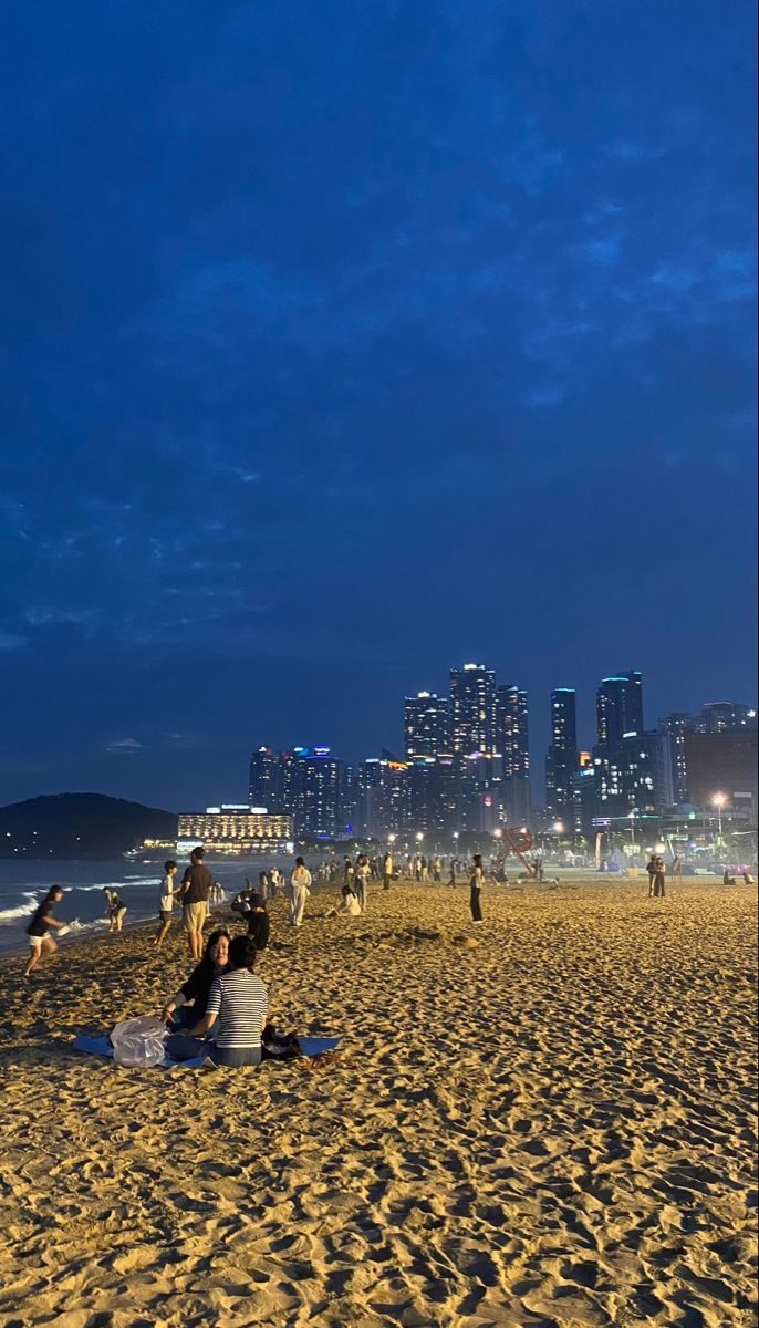 people sitting on the beach at night with city lights in the backgroung