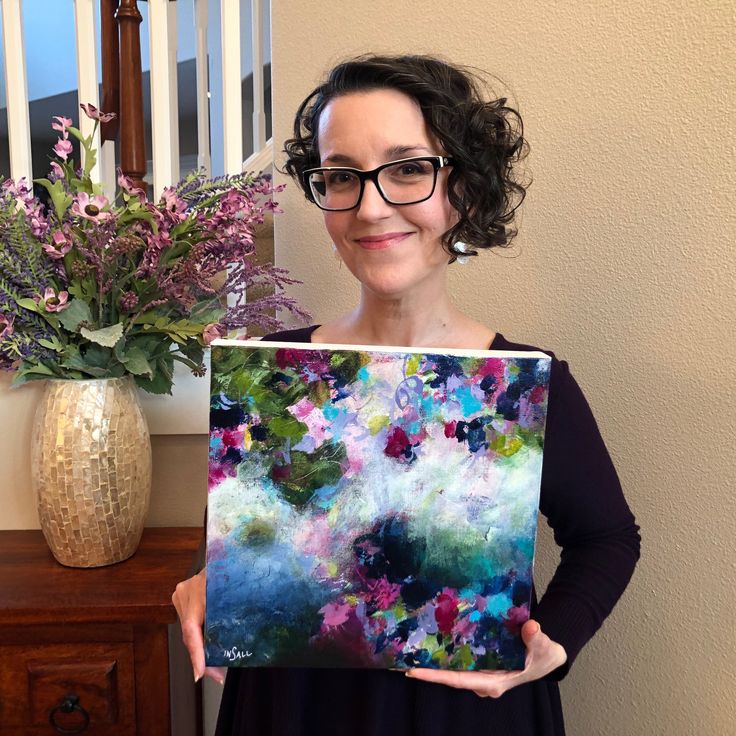 a woman holding up a painting in front of a vase with purple flowers on it