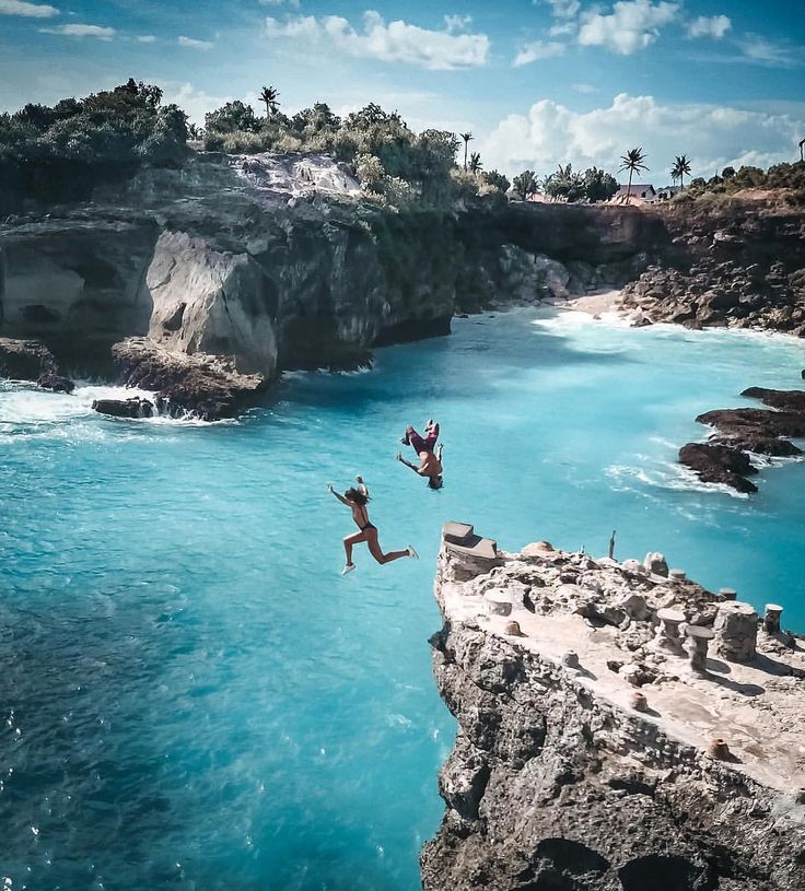 two people jumping off rocks into the ocean with blue water and cliffs in the background