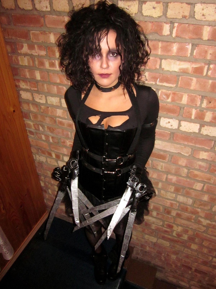 a woman dressed in black and holding two large scissors next to a red brick wall