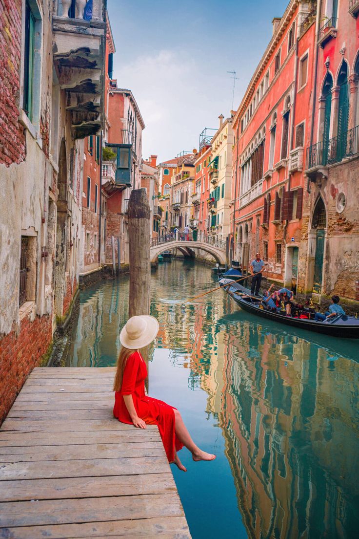 a woman in a red dress and hat sitting on a dock next to a canal