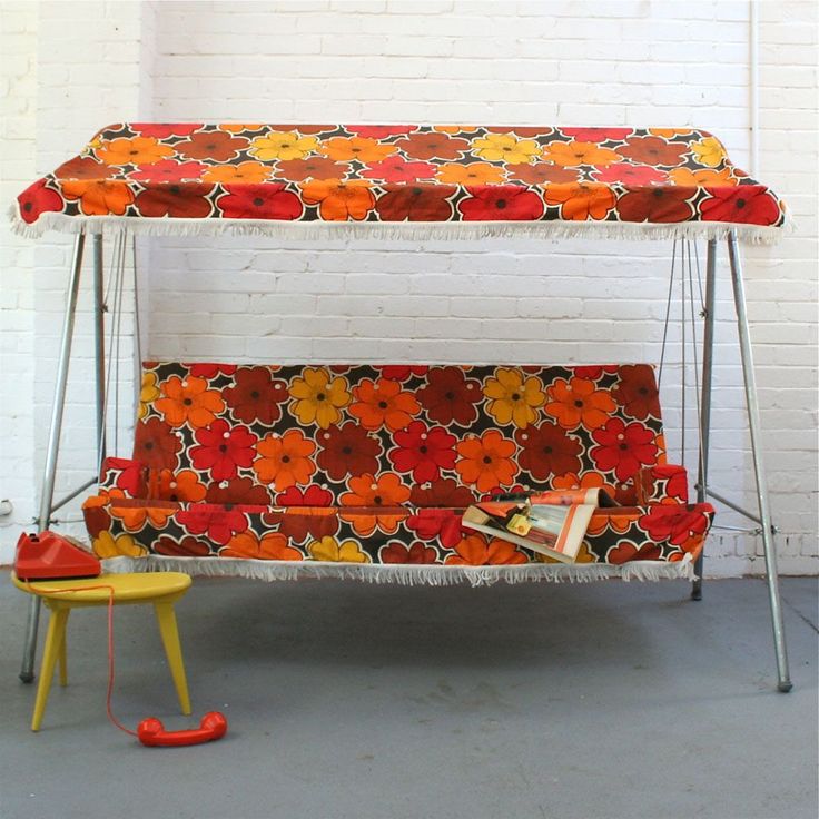 an orange and red bench with flowers on it next to a yellow chair in front of a white brick wall