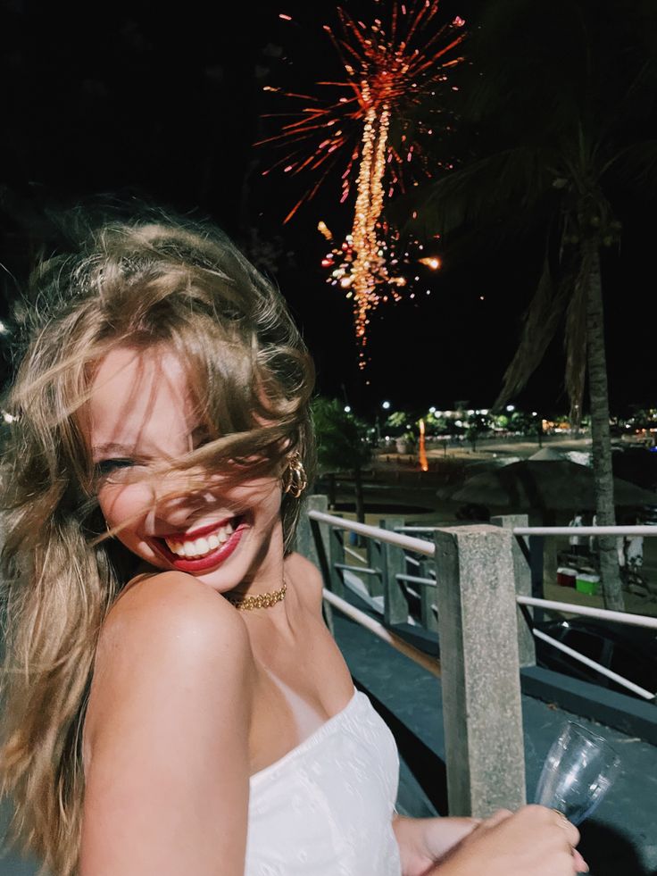 a woman in white shirt standing next to a fence with firework behind her and smiling at the camera