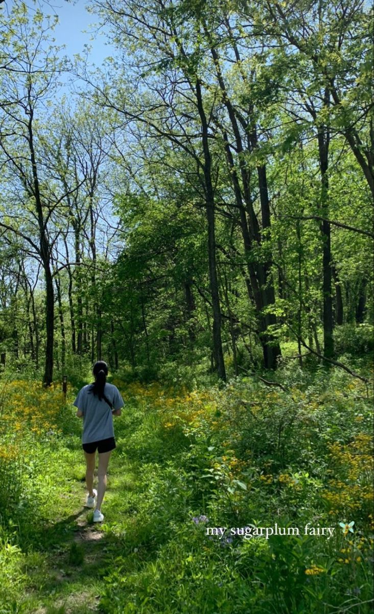 a woman walking through a lush green forest filled with lots of trees and wildflowers