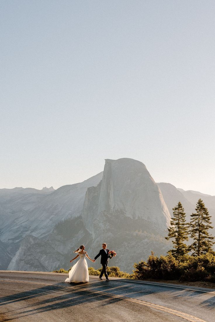 a bride and groom walking down the road in front of a mountain range at sunset