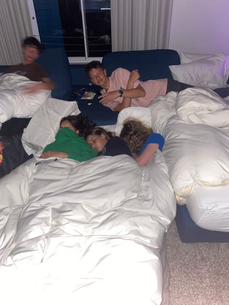 four people laying on a bed in a room with white sheets and pillows, all looking at the camera