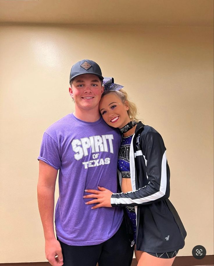 a man standing next to a woman in a purple shirt and black shorts with her arms around her chest