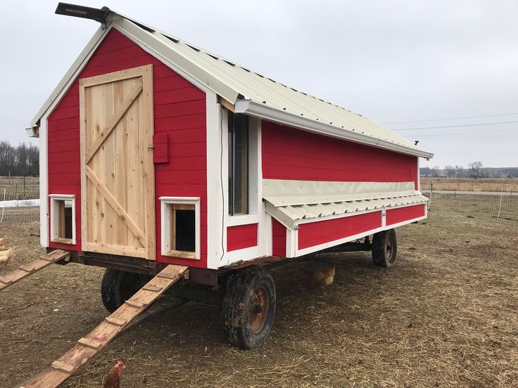 a small red and white chicken coop on wheels in a field with an open door