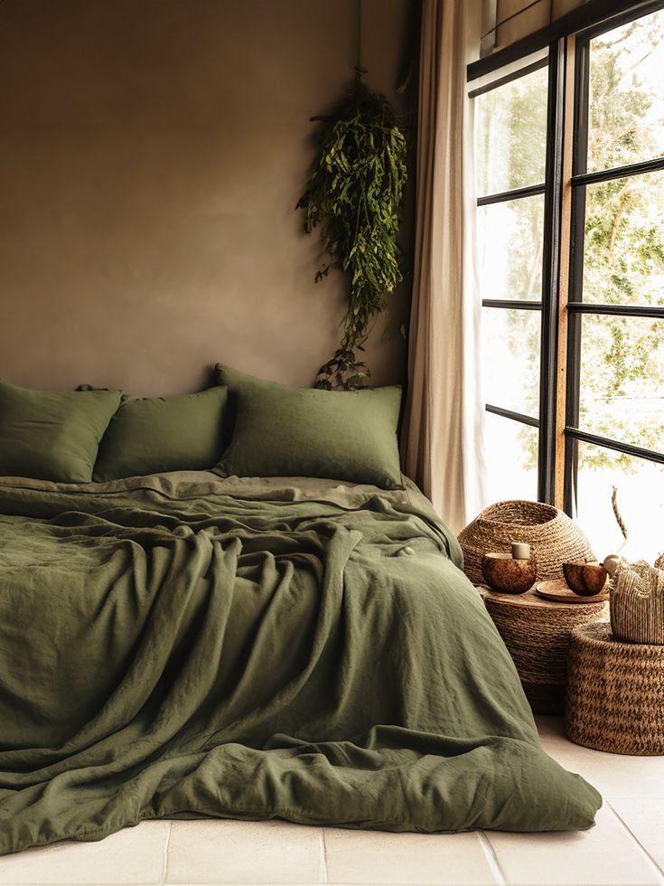 a bed with green sheets and pillows in front of a window next to a basket