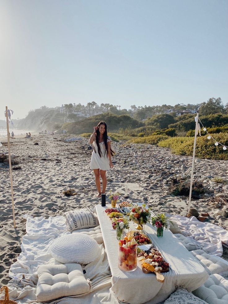 a woman standing next to a table with food and drinks on it at the beach