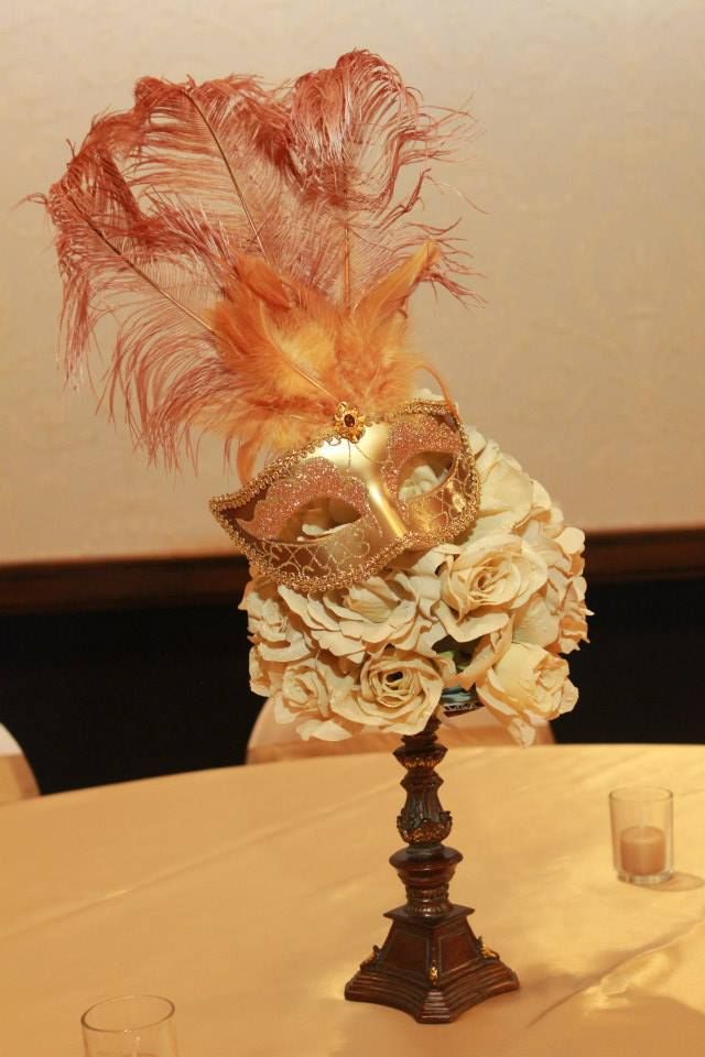 a table topped with a tall vase filled with flowers and feather covered masks on top of it