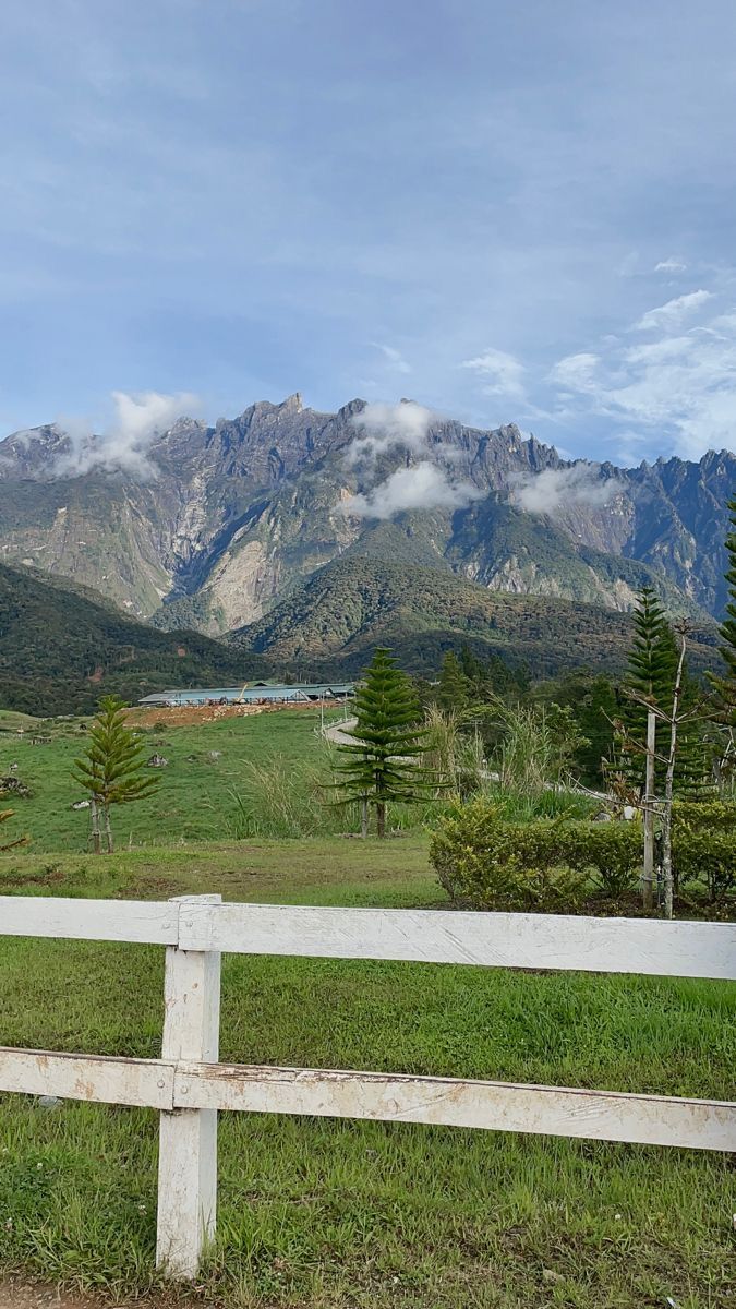 a white fence in front of mountains with trees and grass on the ground below it