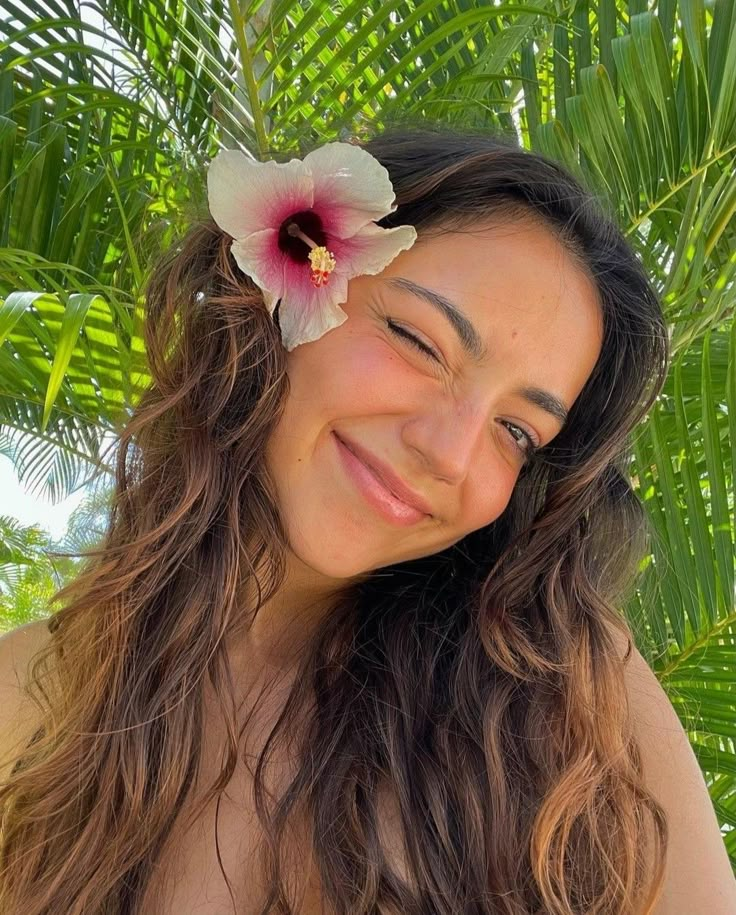 a woman with long hair and a flower in her hair smiles at the camera while standing under palm trees