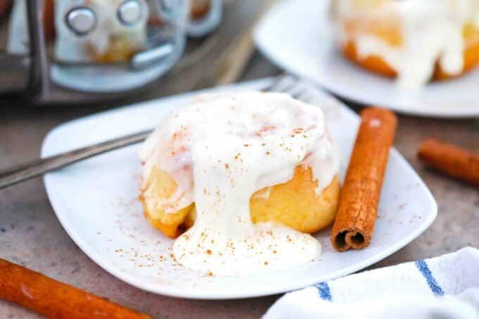 cinnamon buns with icing on white plates next to cinnamon sticks and coffee pot