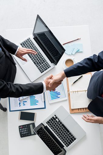 two people shaking hands over a table with laptops