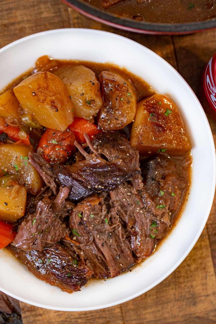 a white bowl filled with beef and potatoes on top of a wooden table next to a can of coke