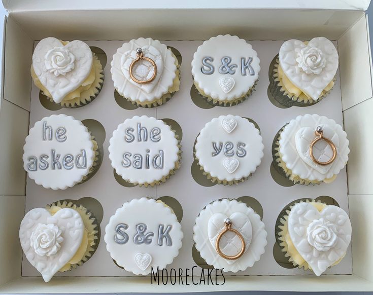 twelve cupcakes in a box decorated with wedding rings and engagement rings, the words she asked, she said, s & k