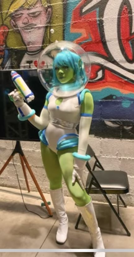 a woman dressed as an alien holding a paint roller in front of a wall with graffiti on it