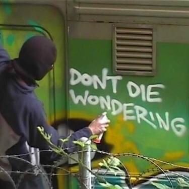 a man in a black hoodie writing on a green wall next to barbed wire