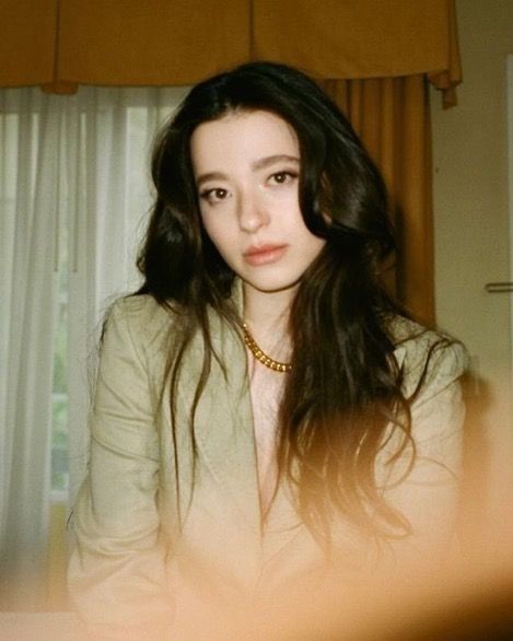 a woman with long dark hair wearing a white shirt and gold necklace sitting in front of a window