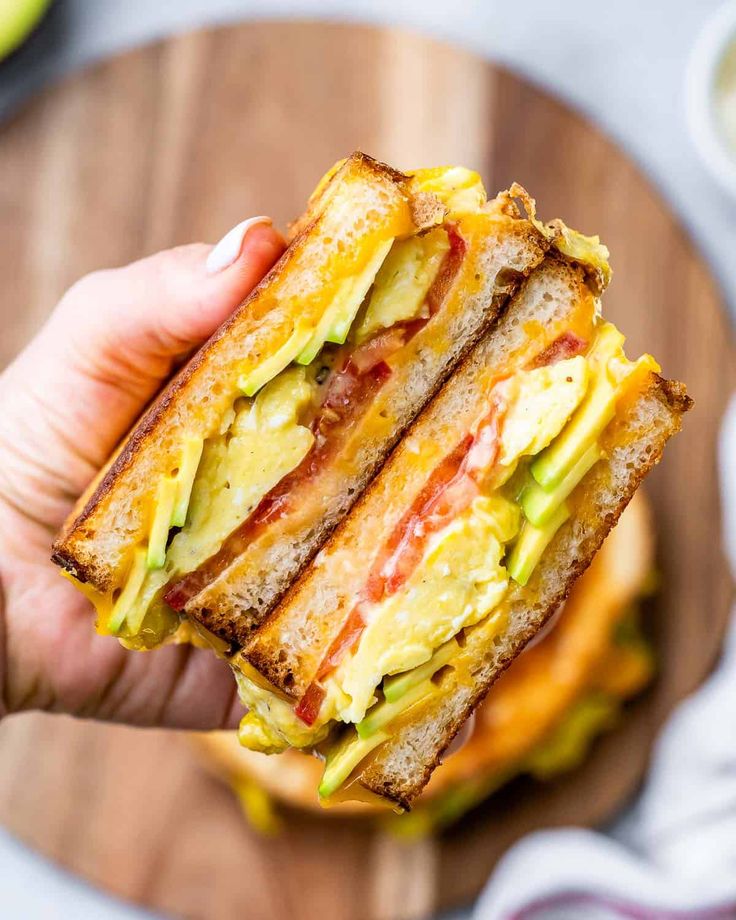 a person holding up a sandwich with eggs and avocado in the middle on a cutting board