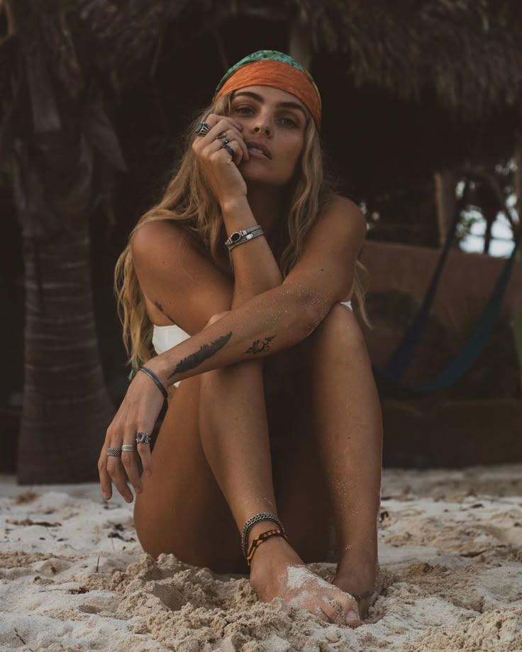 a woman sitting in the sand with her hand on her face and wearing a bandana