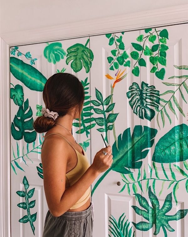 a woman standing in front of a wall with green leaves on it and painting the walls