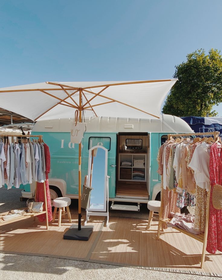 an outdoor market with clothes on racks and umbrellas over the door, in front of a van