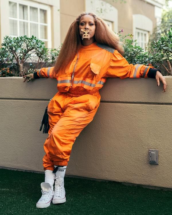 a woman in an orange jumpsuit leaning against a wall with her hands on her hips