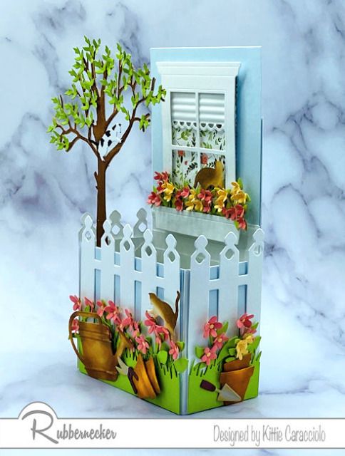 an open card with a tree and flowers in the window sill next to a white picket fence