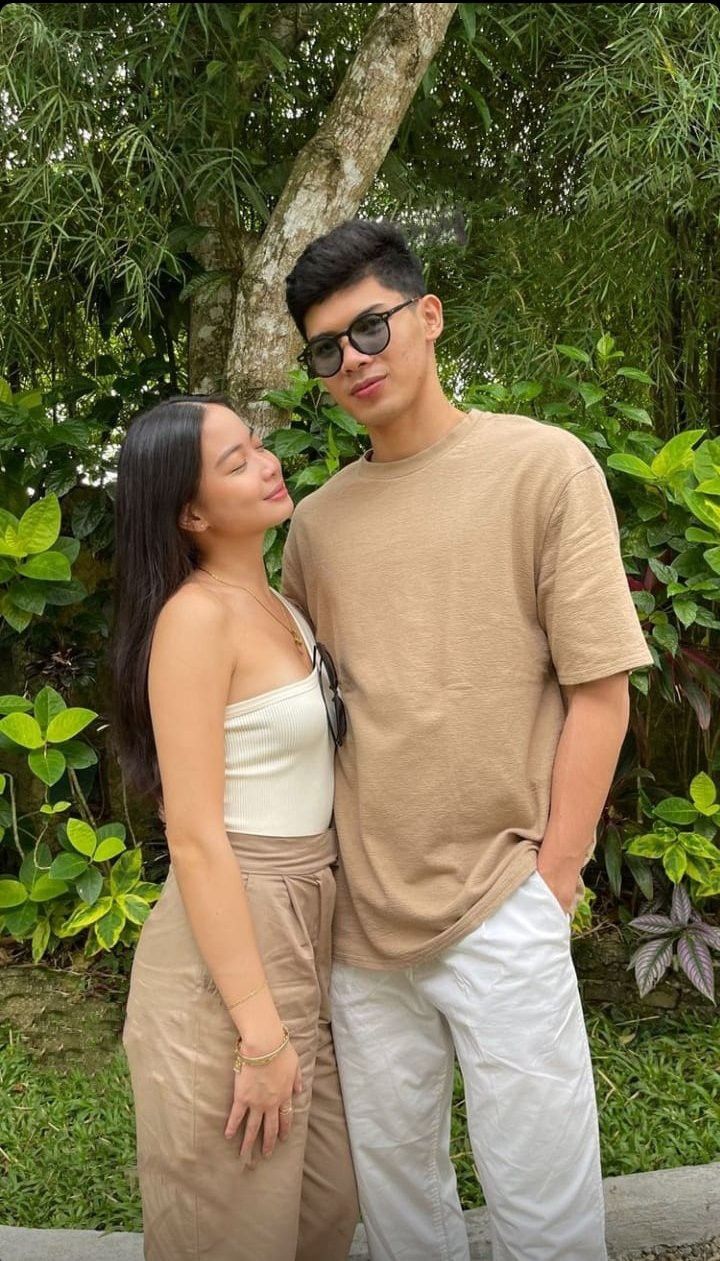 a man and woman standing next to each other in front of some bushes with trees behind them